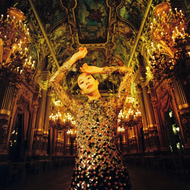 Elegant Asian woman in shimmering gold dress poses in an ornate ballroom with chandeliers and ceiling art. Fashion, glamour, style.