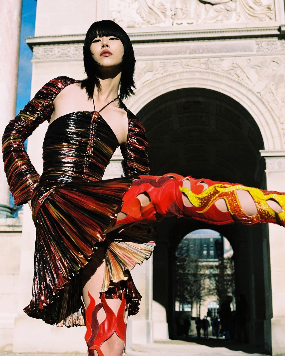 A stylish Asian woman in a shimmering dress poses beneath an ornate stone arch, showcasing bold, flame-inspired thigh-high boots.