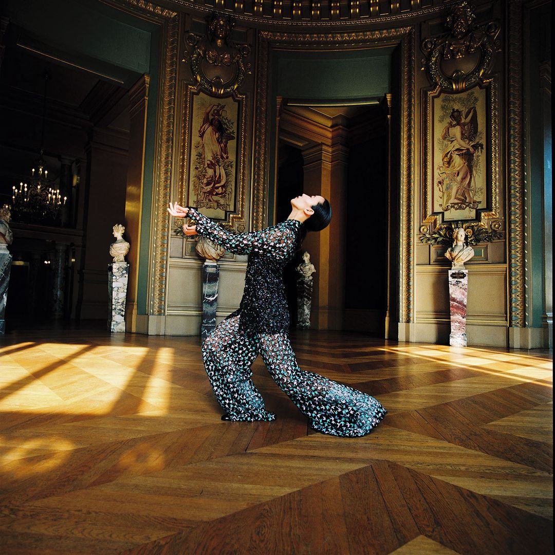 Fashion photography in an ornate hall. A woman poses in a patterned, sheer outfit, with a parquet floor and detailed wall art in background.
