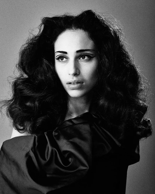 Black and white portrait of a woman with dark wavy hair, wearing dark draped garment, and dark eyeliner, against a grey background.