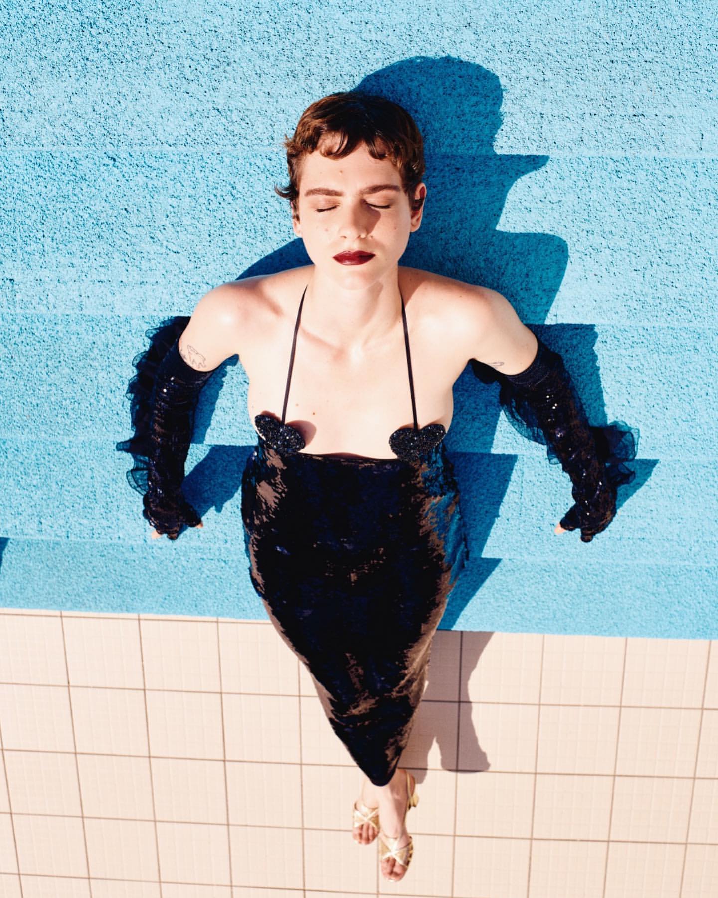 Woman in sequin dress poses on blue poolside tile, wearing gloves and sandals, eyes closed in bright sunlight, overhead shot.