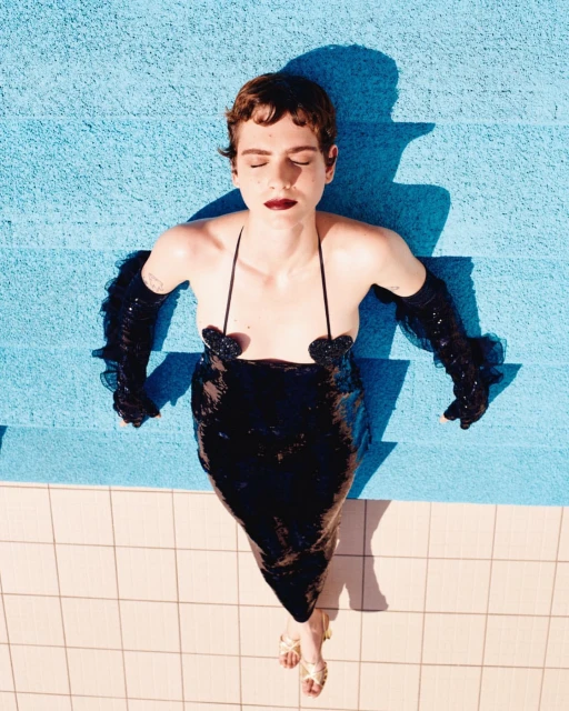 Woman in sequin dress poses on blue poolside tile, wearing gloves and sandals, eyes closed in bright sunlight, overhead shot.