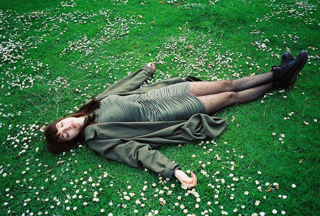 Young woman in olive dress and coat rests on grassy field with daisies. Her legs are covered in black dotted tights and black boots.