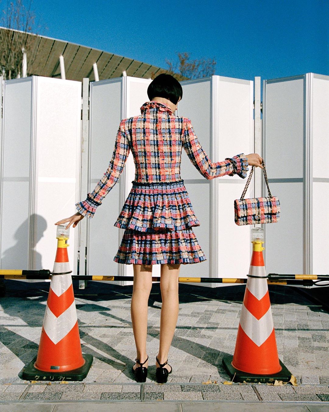 Fashion shot: Woman in a plaid jacket and skirt stands between orange traffic cones against a bright blue sky with panels behind.