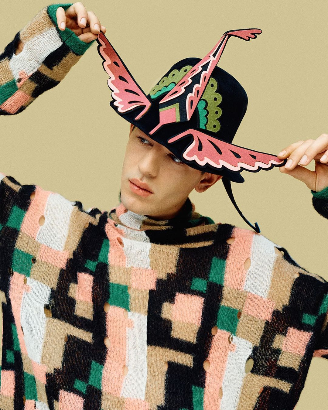Adult male wearing a geometric patterned sweater and a black hat with butterfly-wing decoration against a tan background.