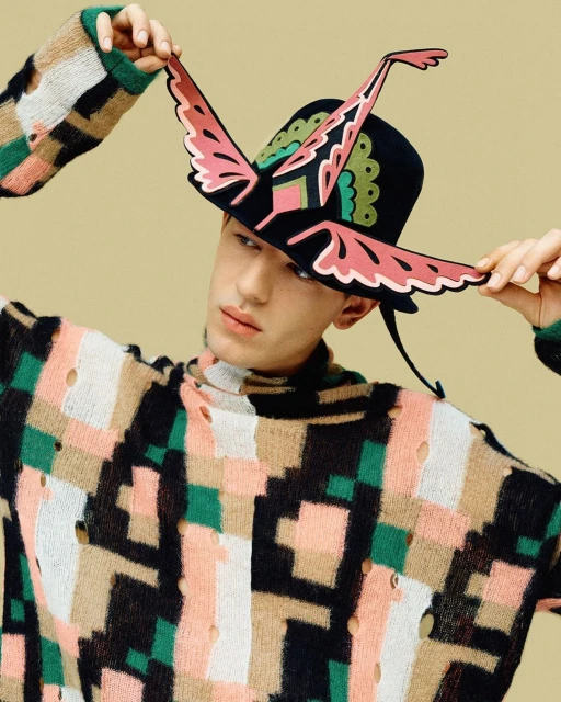 Adult male wearing a geometric patterned sweater and a black hat with butterfly-wing decoration against a tan background.