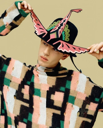 Adult male wearing a geometric patterned sweater and a black hat with butterfly-wing decoration against a tan background.