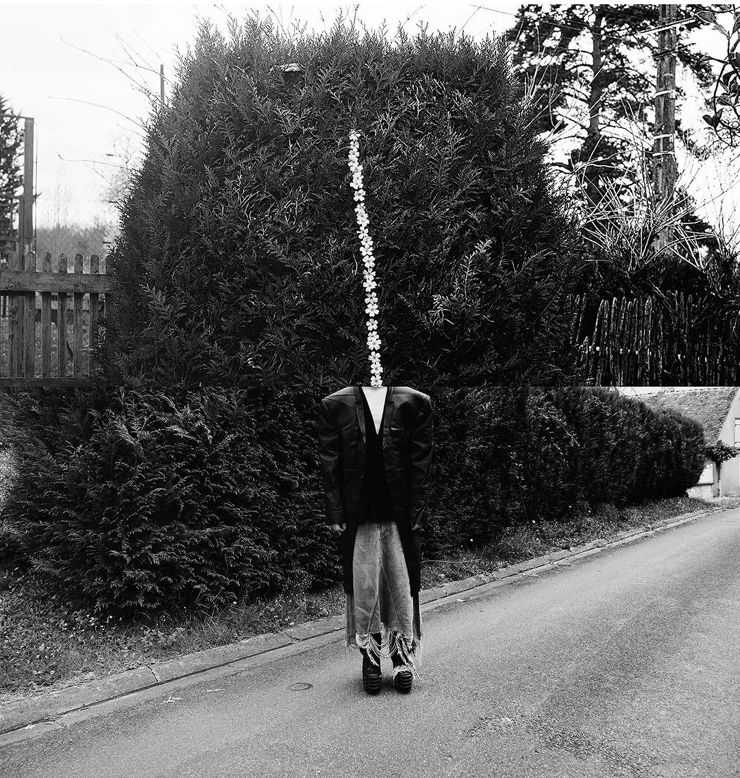 Surreal black-and-white outdoor shot of a person in a suit standing on a road, with a head replaced by a string of flowers. 