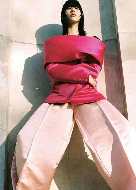 Woman with dark hair poses in pink top and light pants against a concrete wall, highlighted by natural light and deep shadows.
