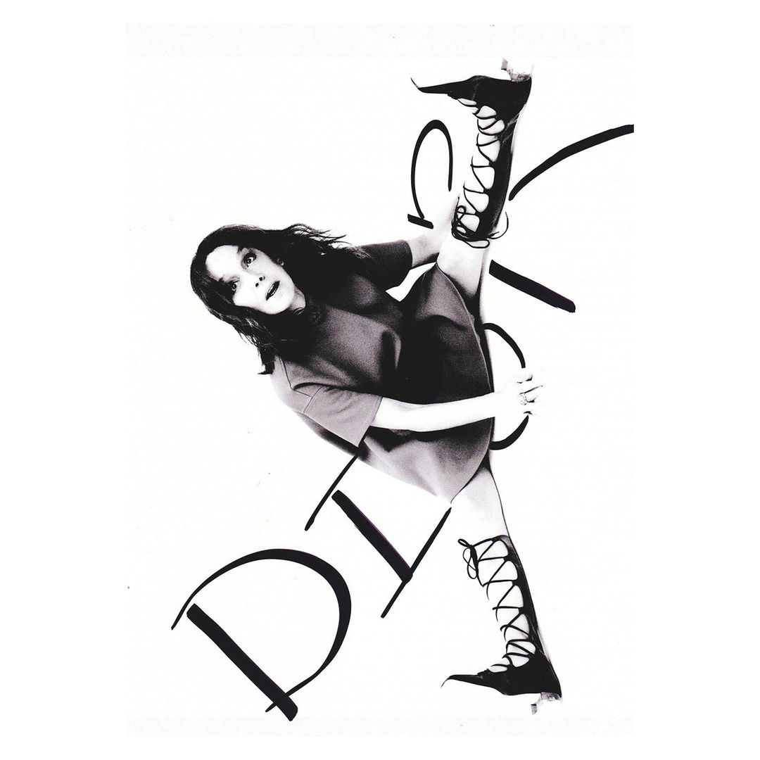 A woman with dark hair and a dress, performs a leg split in lace-up boots, posing against a white background with large black handwritten text.
