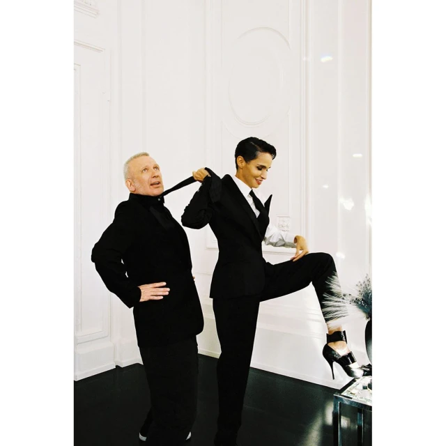 An older man and a younger woman pose in black suits against a white wall, the woman holding the man's tie, with a glossy floor.