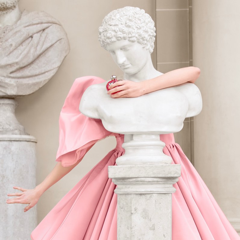 Bust statue holds pink perfume bottle, contrasting with a pink dress. Another bust is visible in background. Soft, elegant studio style.