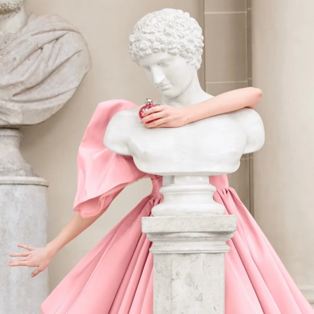 Bust statue holds pink perfume bottle, contrasting with a pink dress. Another bust is visible in background. Soft, elegant studio style.