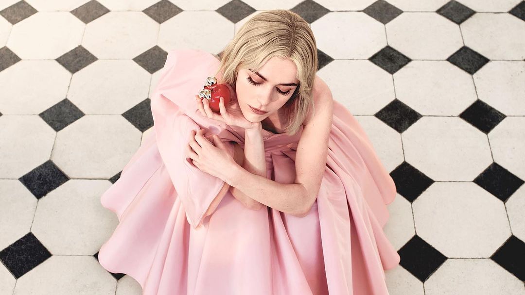 Adult woman in a pink gown poses indoors with a red decorative apple, set against a black and white tiled floor in a portrait.