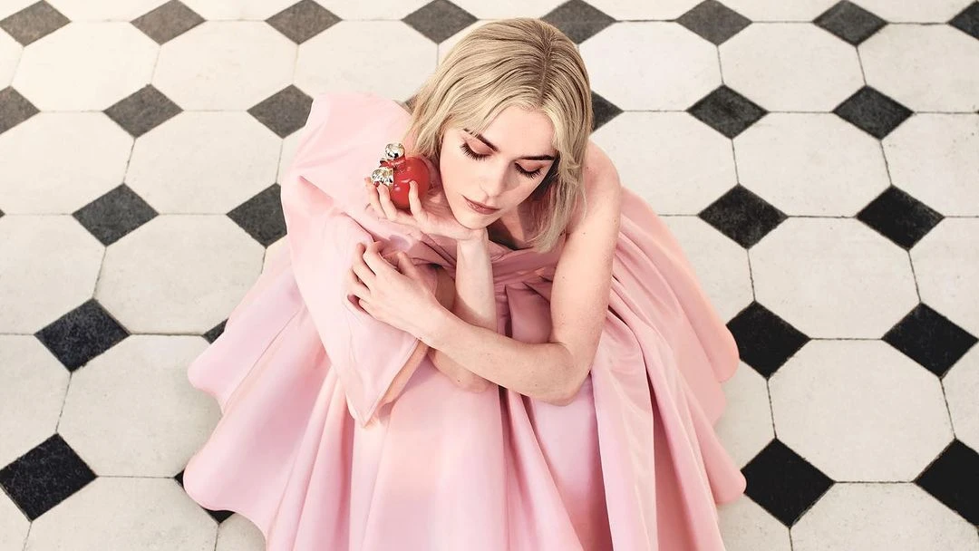 Adult woman in a pink gown poses indoors with a red decorative apple, set against a black and white tiled floor in a portrait.