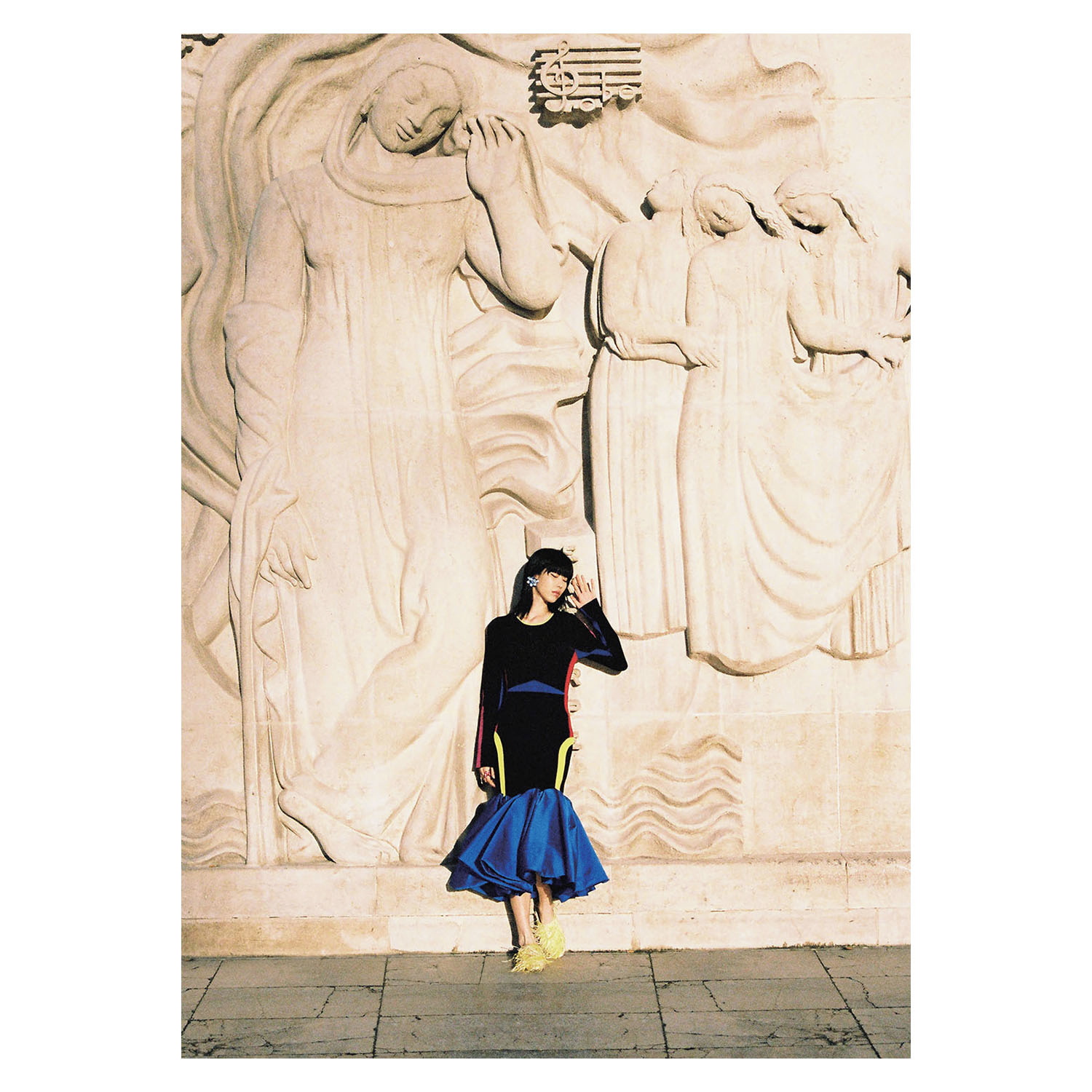 Adult female in a black and blue dress poses in front of a stone carving with musical notes and figures in an outdoor setting.