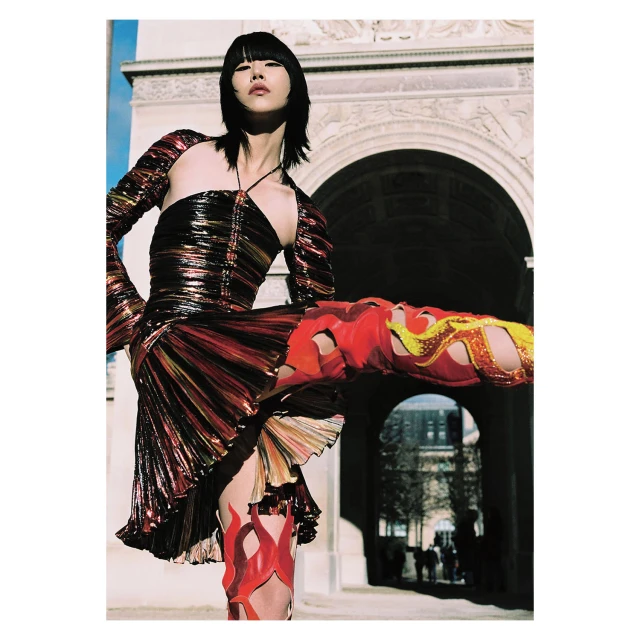 Stylish young woman in a metallic dress and flame-patterned boots poses in front of a Parisian archway on a bright day.