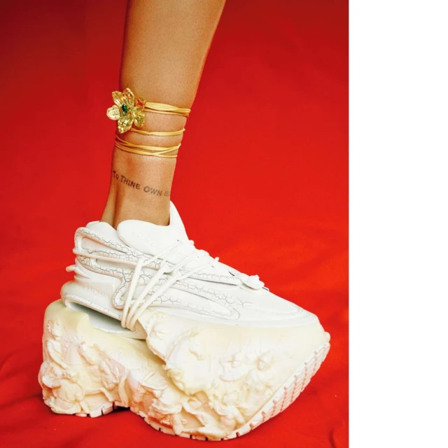 Elevated fashion: white platform sneaker on red. Gold ankle jewelry and tattoo detail. Focus on footwear and ankle adornments.