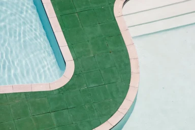 High-angle view of a swimming pool edge featuring green square tiles, white coping stones, and clear light blue water.