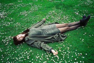 Young woman in an olive green coat and dress lying flat on a vibrant green lawn scattered with tiny white flowers