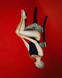 A bald woman in a black swimsuit perched on a patterned stool against a vivid red background