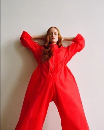 Redheaded model wearing a vibrant red jumpsuit posing with arms behind head against a plain off-white wall
