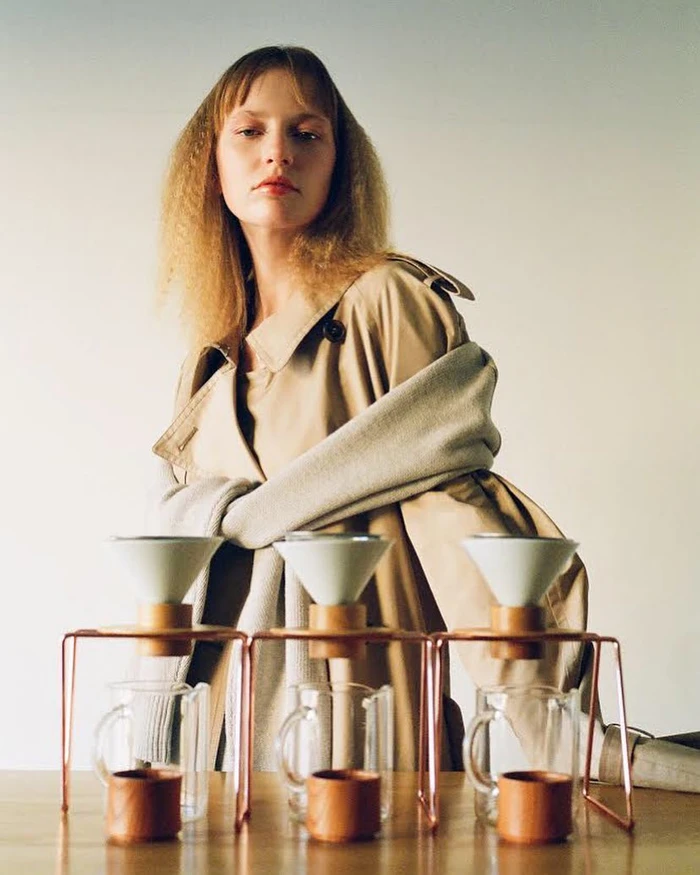 Woman in a beige trench coat standing behind three pour-over coffee drippers on a wooden table by The Bardos