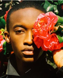Close-up portrait of a young Black man holding bright red flowers up to the side of his face amongst green foliage.