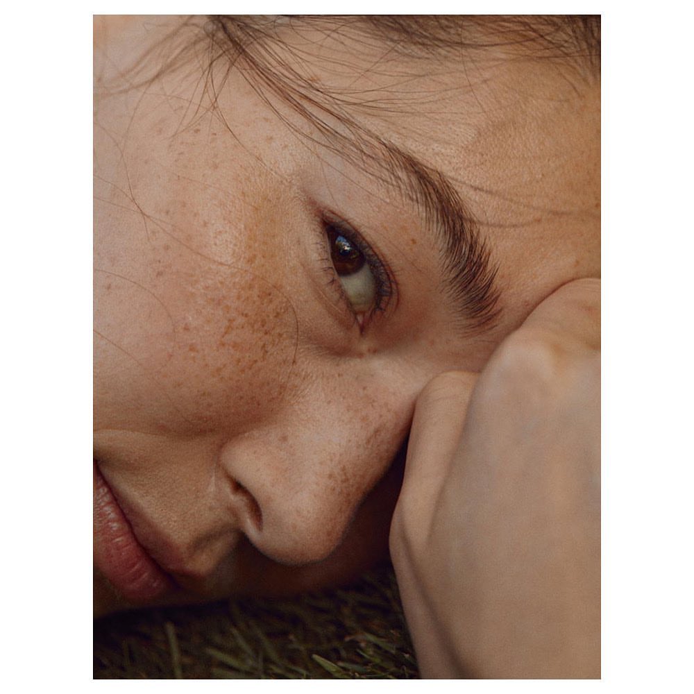 Close-up portrait of a young adult female with freckles resting her head on her hand in an outdoor setting with neutral color palette.