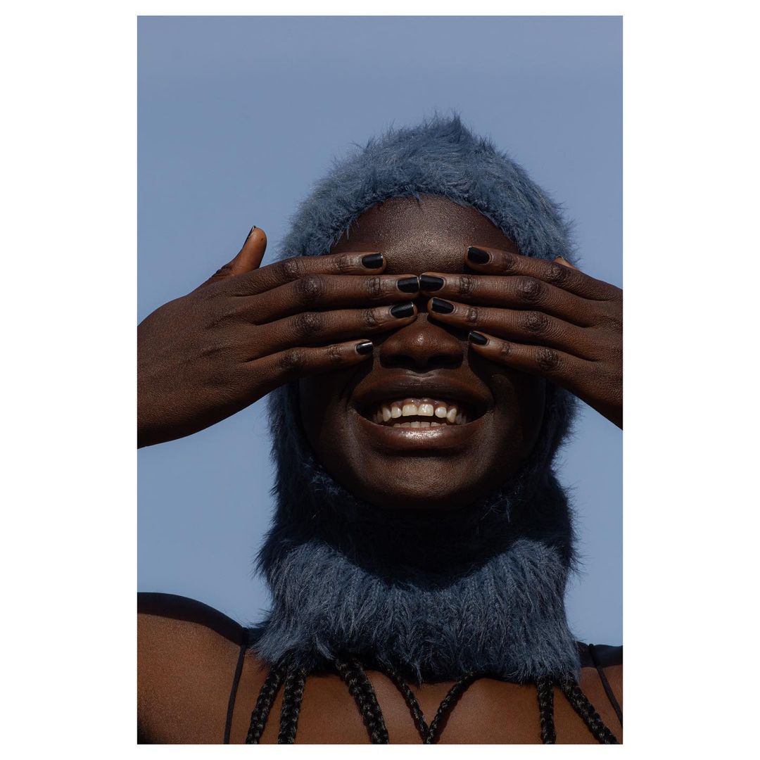 Close-up of an adult woman with dark skin wearing a fuzzy blue balaclava covering her hair, with both hands covering her eyes, smiling.