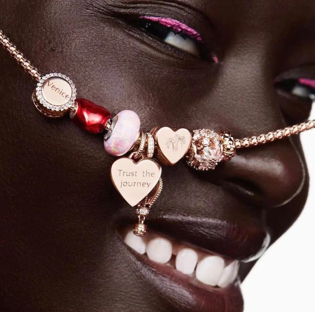 Close up of a Black woman smiling, adorned with rose gold Pandora bracelet featuring charms. Bracelet drapes from above her nose to upper lip.
