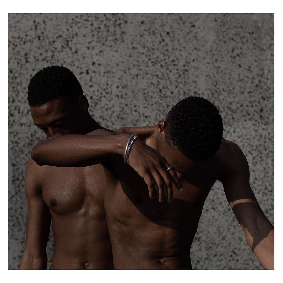 Striking studio portrait of two dark-skinned men with bare chests, close together against a textured grey backdrop, showcasing their skin tones.