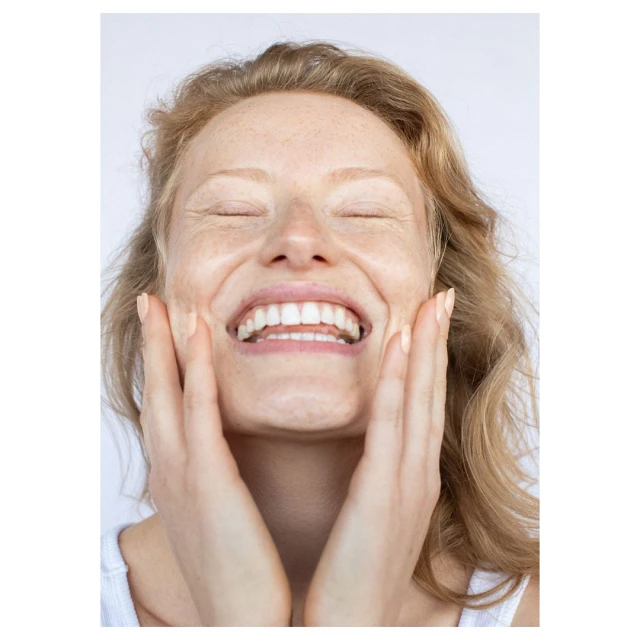 Close-up portrait of a smiling woman with hands on her cheeks in a bright, minimalist setting, showcasing skincare or beauty product effects.