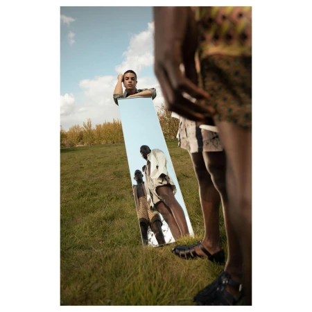 Man holding a tall vertical mirror standing in a grassy field reflecting another person outdoors