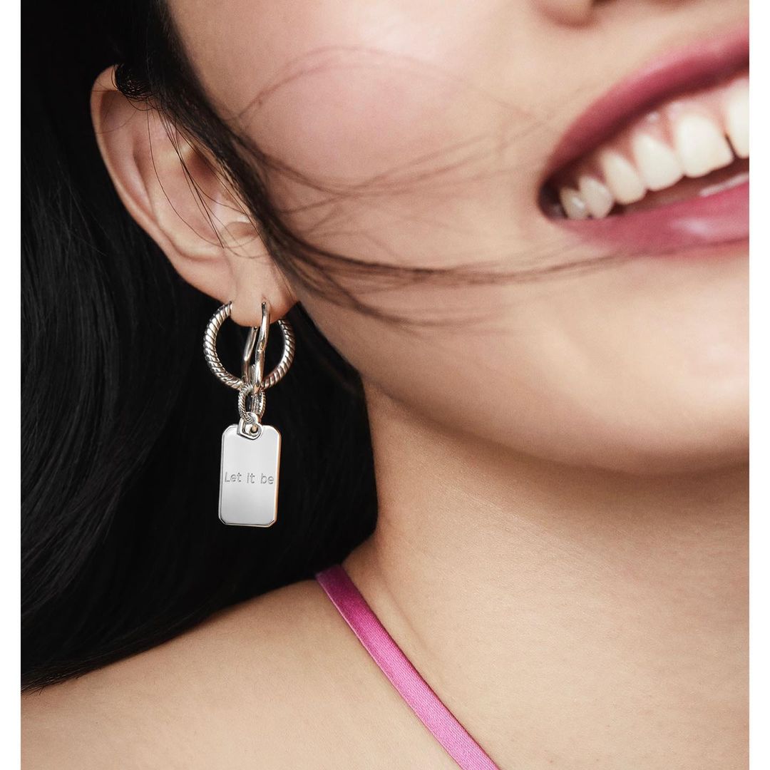 Close-up portrait of a woman wearing a silver hoop earring and a tag pendant, smiling with pink lipstick.
