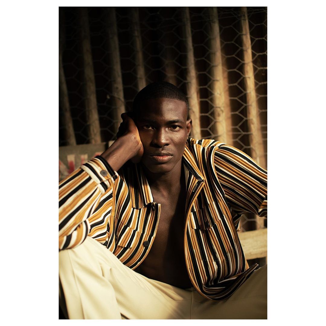 Portrait of an African American male in a striped jacket and tan pants, posed indoors against a textured wall in warm, dramatic lighting.