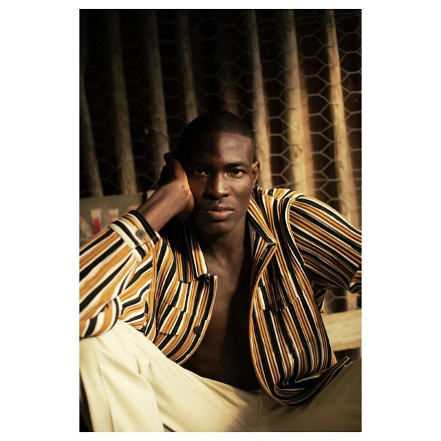 Portrait of an African American male in a striped jacket and tan pants, posed indoors against a textured wall in warm, dramatic lighting.