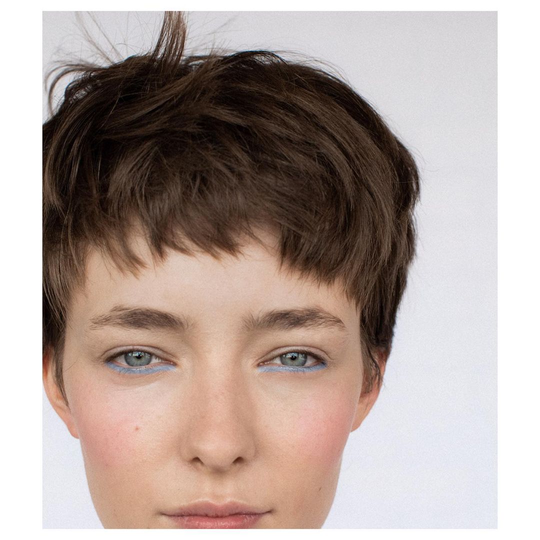 Close-up portrait of a young adult female with short brown hair and blue eyeliner on a white background, showcasing beauty and makeup.