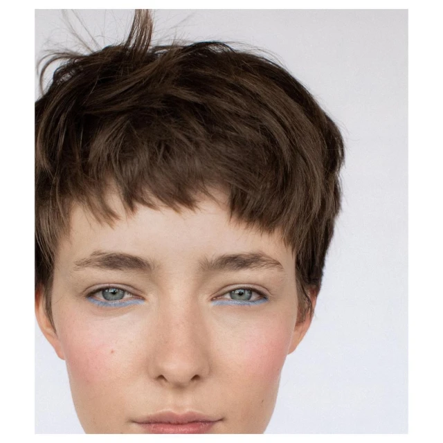 Close-up portrait of a young adult female with short brown hair and blue eyeliner on a white background, showcasing beauty and makeup.