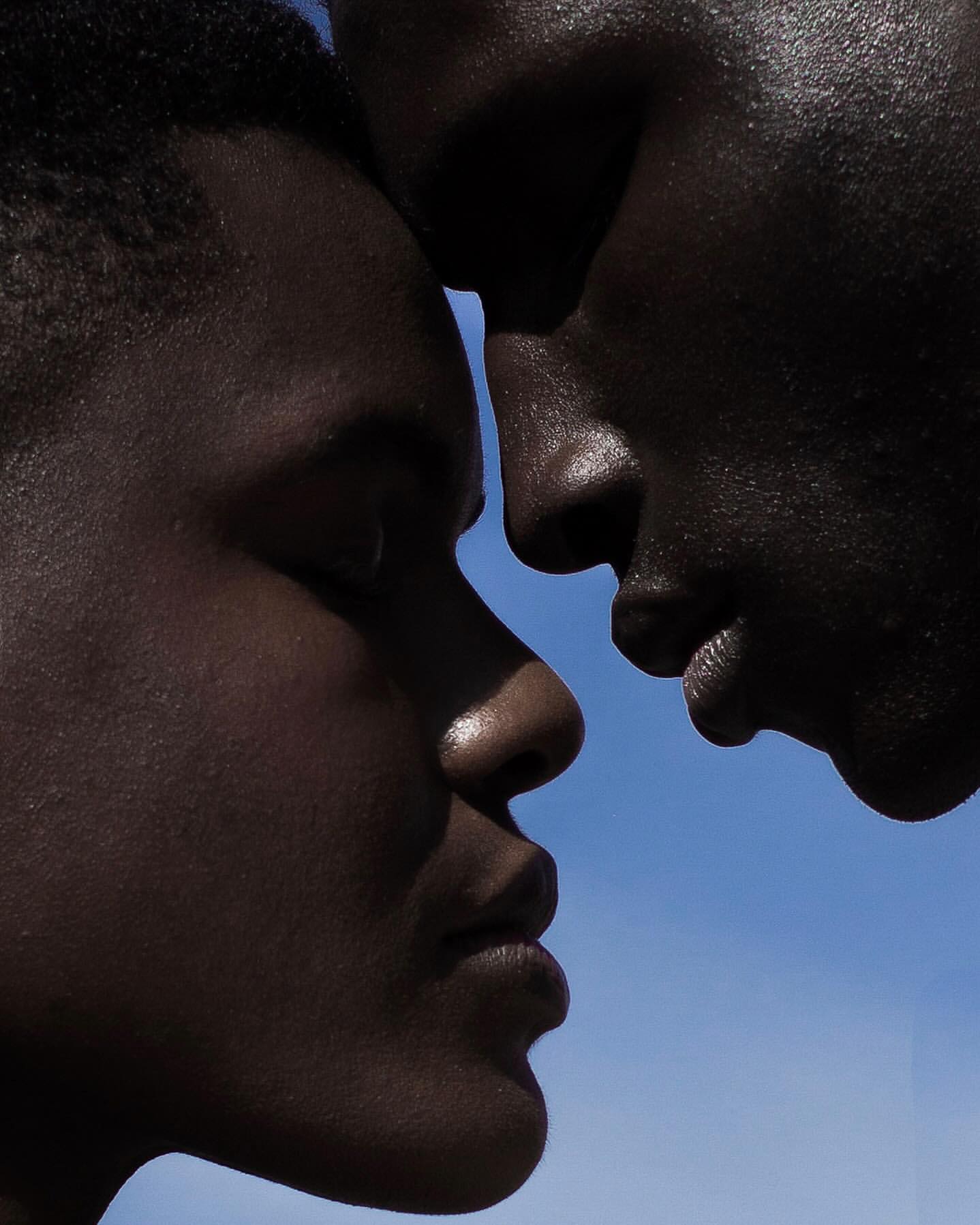 Close-up of two adult humans of African descent standing face-to-face against a bright blue sky in an outdoor setting.