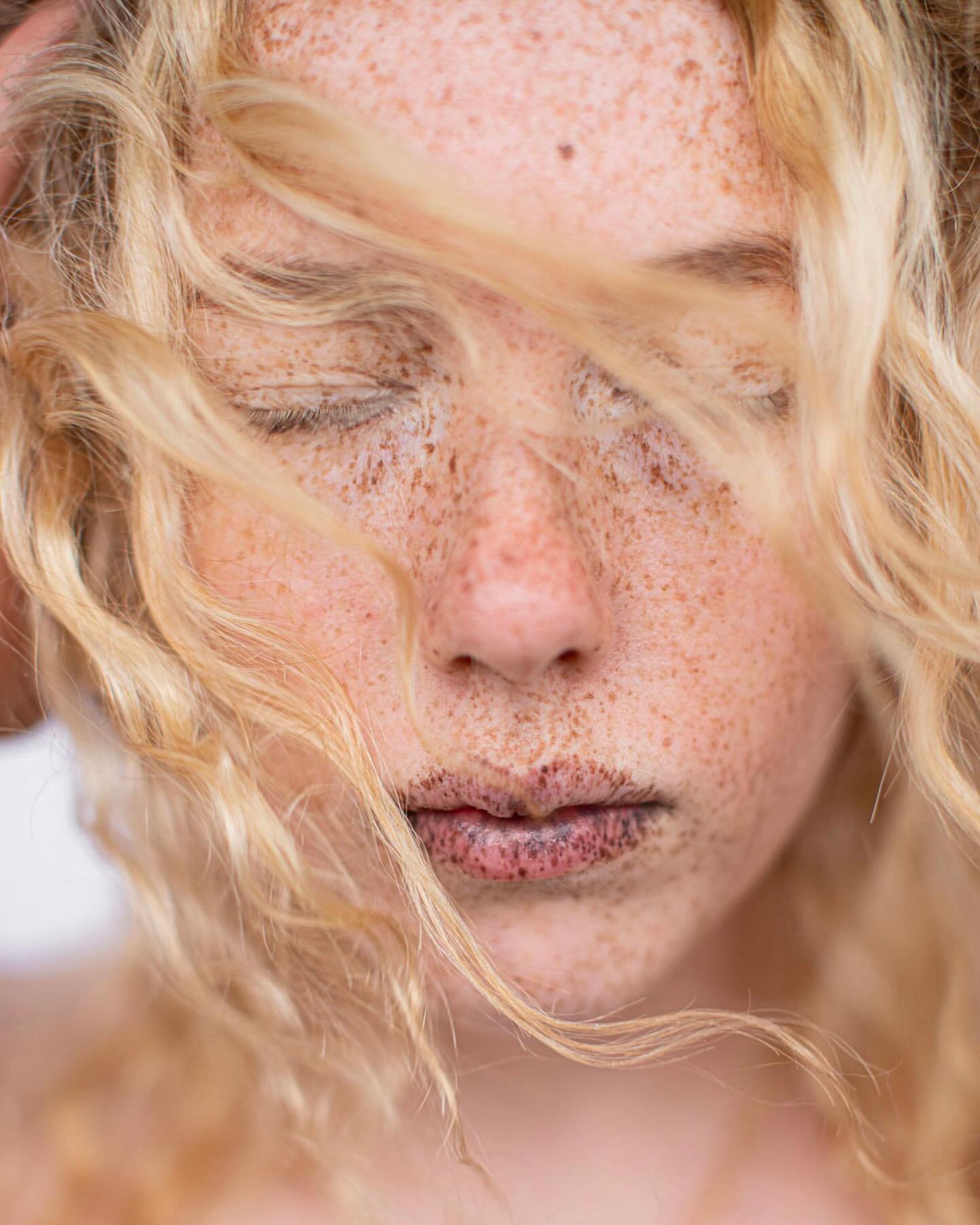 Close-up portrait of a blonde woman with freckles and her eyes closed, showcasing her natural beauty in a soft light setting.