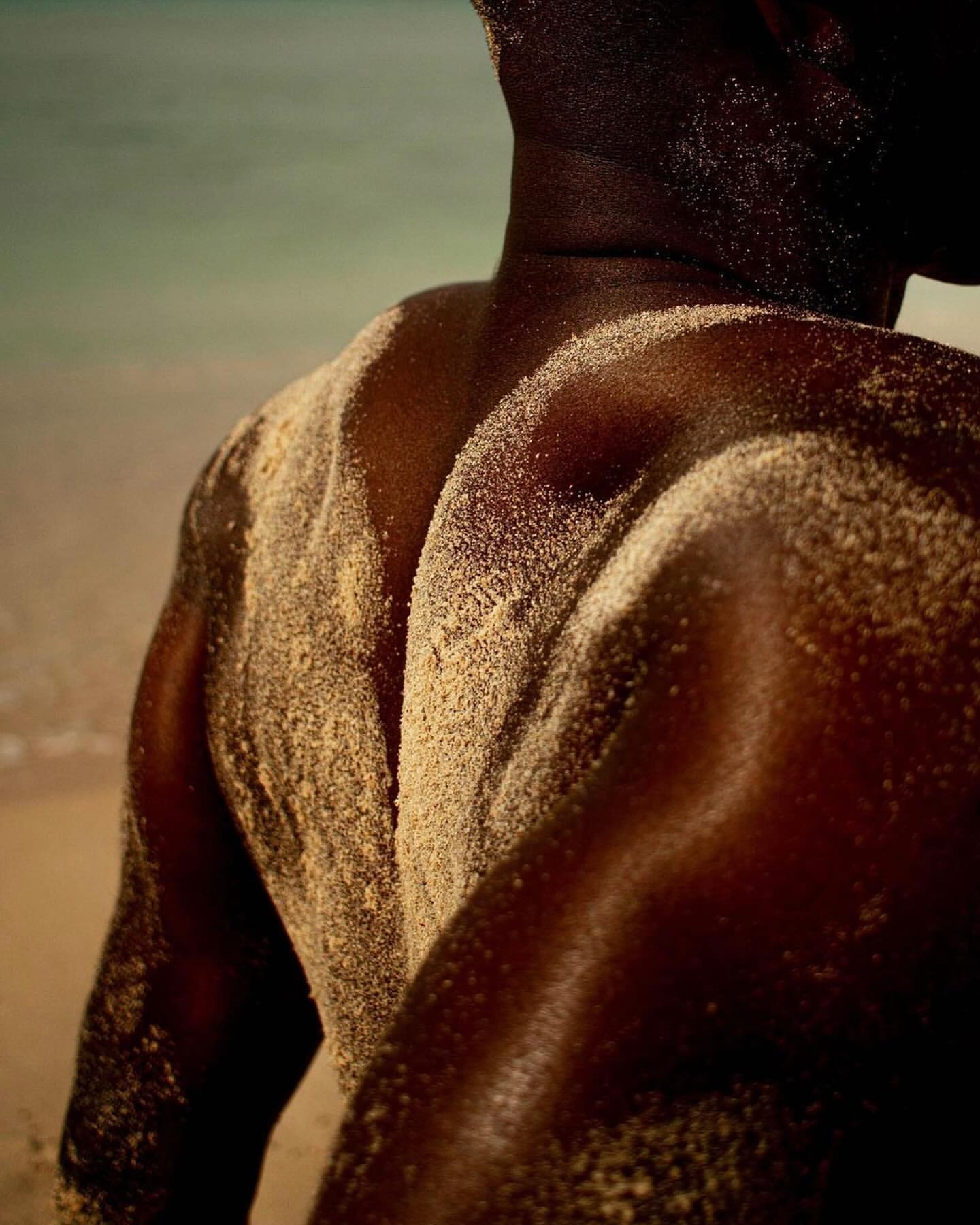 An African male's upper body is dusted with sand on a beach, glistening in the warm sunlight, as waves gently roll in the background.