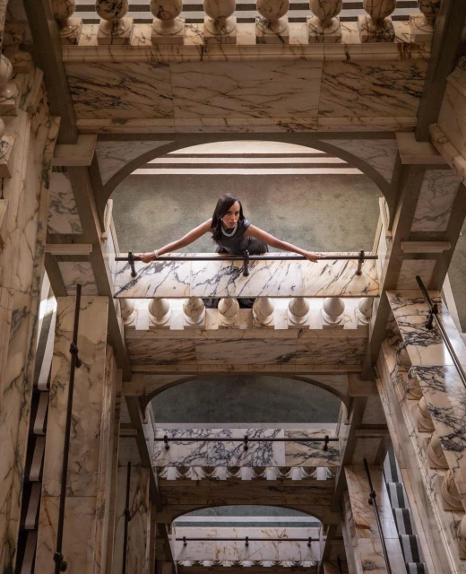 Elegant woman in grey dress leans on marble balcony railing. Indoor shot with architectural detail and neutral, muted color palette.