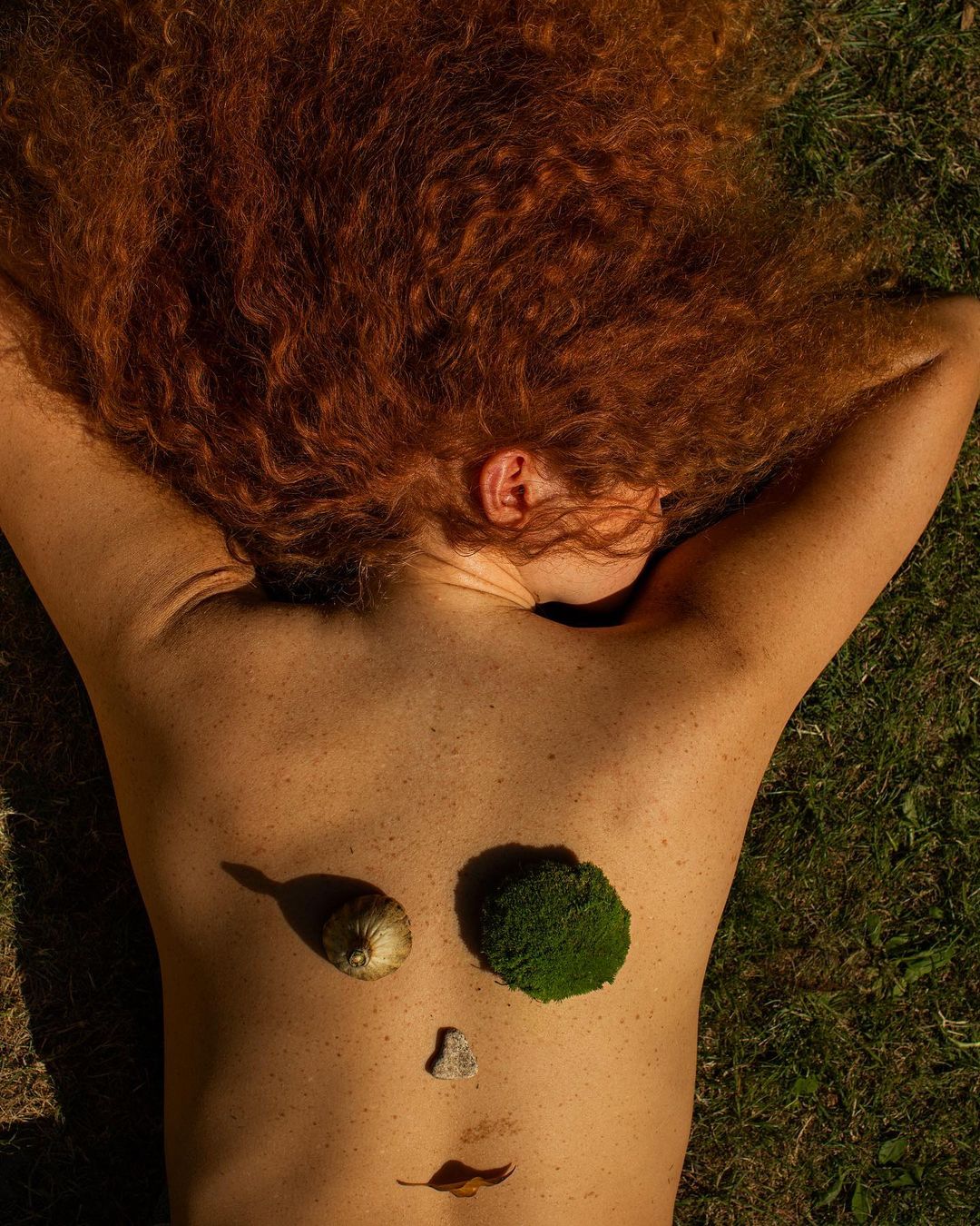 Overhead shot of a woman lying on grass with red hair, face designed with snail shell, moss, stone and leaf in a surreal art photograph.