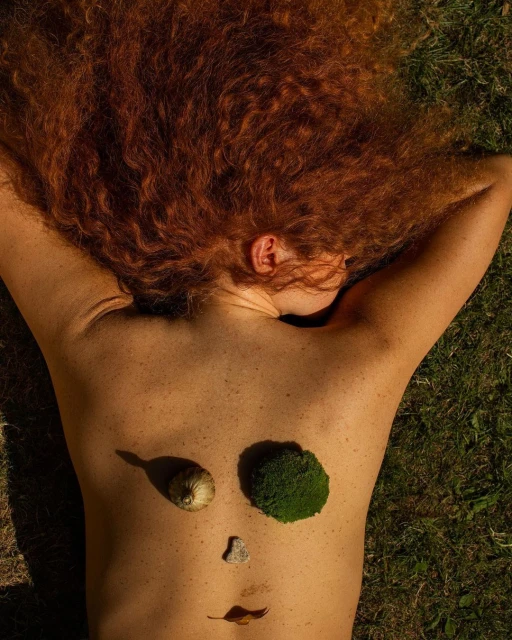 Overhead shot of a woman lying on grass with red hair, face designed with snail shell, moss, stone and leaf in a surreal art photograph.