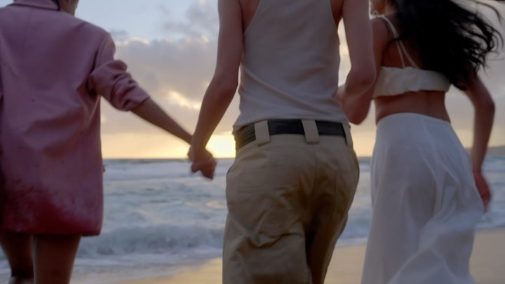 Three people holding hands and running along a sandy beach towards the ocean at sunset.
