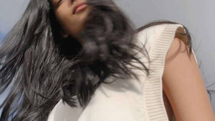 Low-angle shot of a woman with long dark hair wearing a white sleeveless knit top against a clear sky