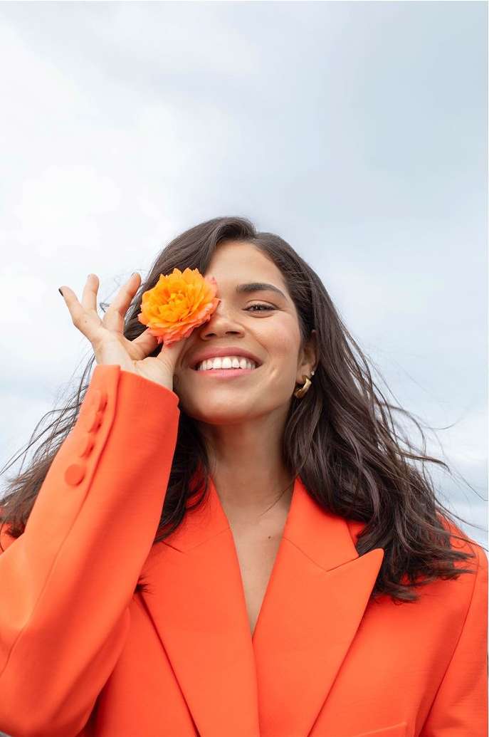 A woman smiles, holding a flower over one eye. She wears a bright orange blazer against a soft sky, creating a cheerful portrait.