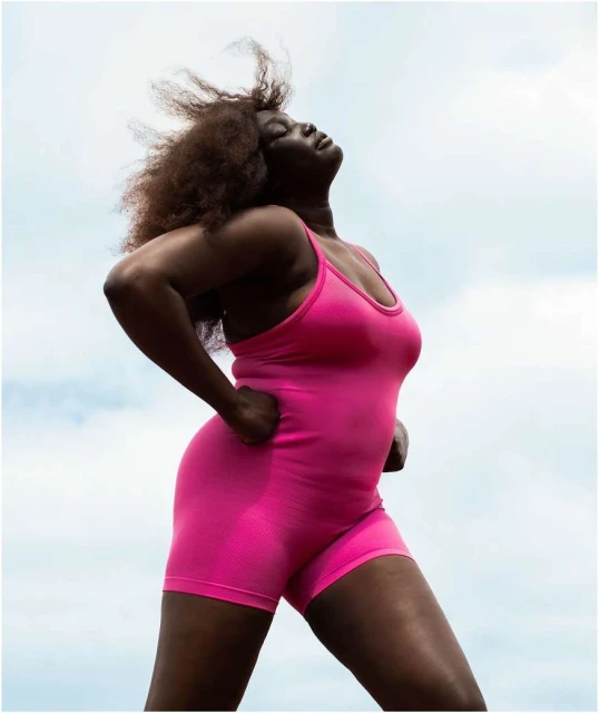 A woman in a pink bodysuit stands outside with her head tilted back and her hands on her hips against a cloudy sky background.