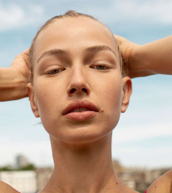 Close-up of a fair-skinned woman with hands behind her head, gazing slightly downward against a backdrop of blue sky and neutral buildings.
